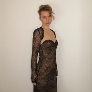 May include: A black lace dress with long sleeves and a sweetheart neckline. The dress is form-fitting and appears to be made of a delicate, floral-patterned lace. The dress is worn over a nude-colored underlay, creating a contrast.