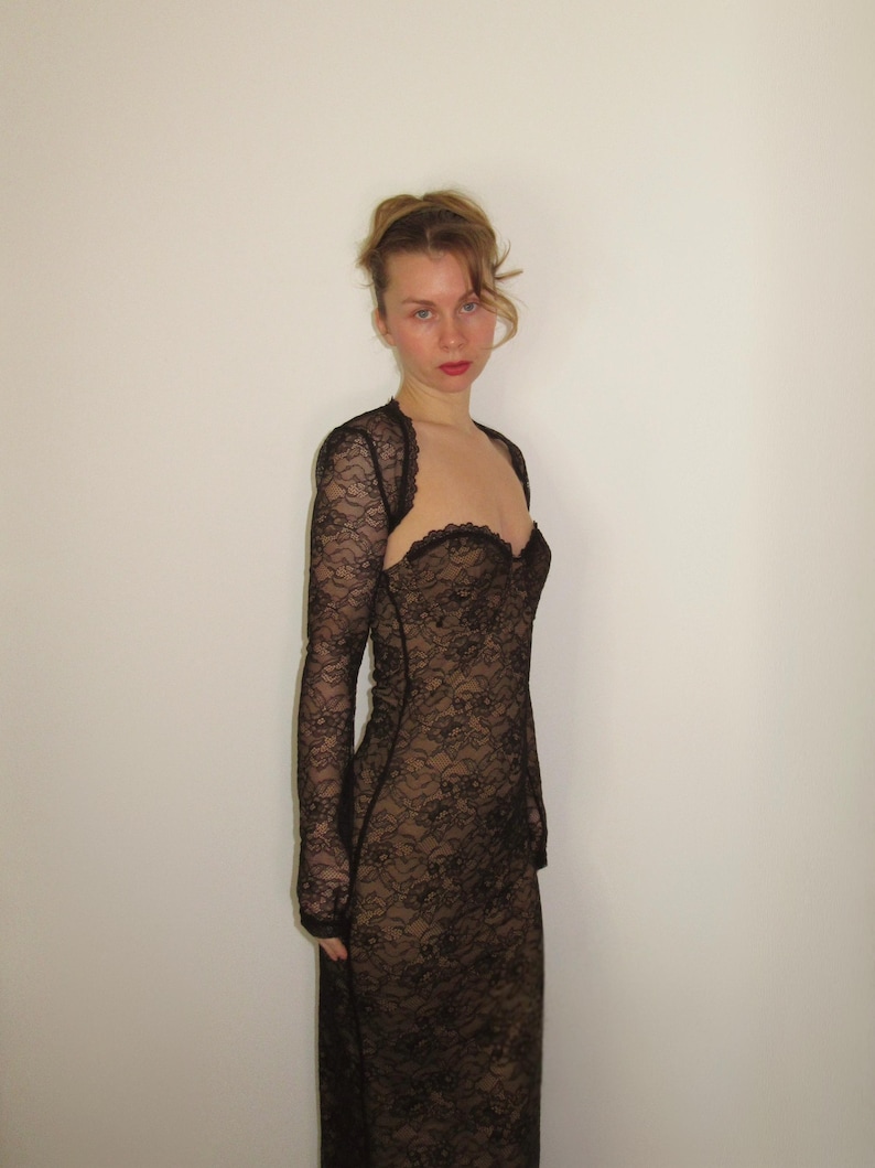 May include: A black lace dress with long sleeves and a sweetheart neckline. The dress is form-fitting and appears to be made of a delicate, floral-patterned lace. The dress is worn over a nude-colored underlay, creating a contrast.