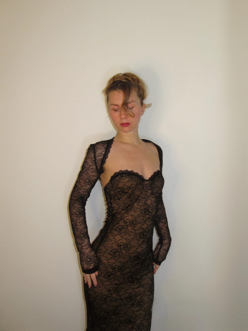 May include: A black lace dress with long sleeves and a sweetheart neckline. The dress is form-fitting and appears to be made of delicate, see-through lace. The sleeves are attached to a bolero-style top.