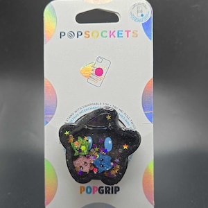 May include: A black star-shaped phone grip with glitter and colourful accents. The grip is on a white card with the text "POPSOCKETS" and "POPGRIP".