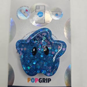 May include: A blue star-shaped phone grip featuring a glittery finish with embedded star and circle sequins. The grip has two black oval eyes and is mounted on a white retail card labeled POPSOCKETS and POPGRIP, made with plant-based materials.
