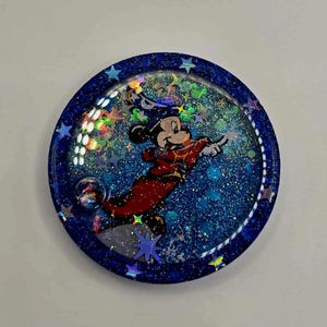 Puede incluir: Un posavasos redondo de resina con purpurina azul que presenta a Mickey Mouse de dibujos animados con un atuendo rojo. El posavasos tiene un borde azul oscuro con detalles en forma de estrella y un interior brillante.