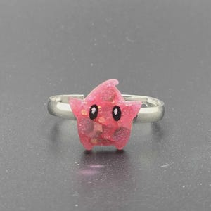 May include: A pink star-shaped ring with a silver band. The star has black eyes and a glittery texture. The ring is a cute and whimsical accessory.