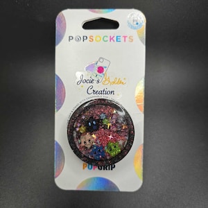 Puede incluir: Un soporte para teléfono PopSockets con un diseño brillante y colorido que presenta pequeñas criaturas de dibujos animados. El agarre es negro con el texto "POPGRIP" en la parte inferior. El embalaje incluye el texto "POPSOCKETS" y "Jocie's Golden Creation".