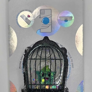May include: A PopSockets PopGrip featuring a green cartoon character inside a black cage design. The packaging includes the PopSockets logo and text indicating it's made with plant-based materials. The PopGrip is designed for phone grip and wireless charging.