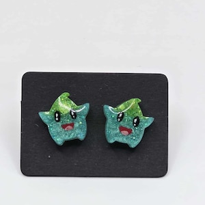 May include: A pair of star-shaped stud earrings. Each earring features a green and blue gradient design with glitter accents, a smiling red mouth, and black eyes. The earrings are displayed on a black card.