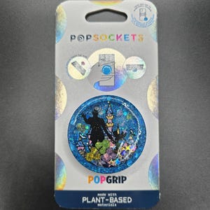 Puede incluir: Un PopSockets PopGrip con un diseño de purpurina azul, con la silueta de un castillo y una figura. El embalaje muestra "POPSOCKETS" y "POPGRI", con el texto "made with PLANT-BASED materials".