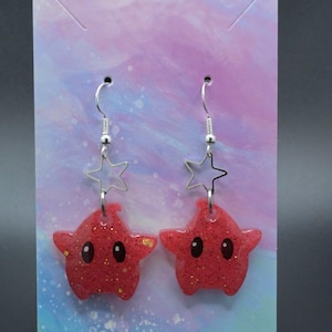 May include: A pair of red star-shaped earrings with glitter and black eyes. The earrings are hanging from silver hooks.