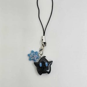 May include: A black charm with blue eyes and a star-shaped charm with blue glitter. The charms are attached to a silver-colored clasp and a black cord. The background is white.