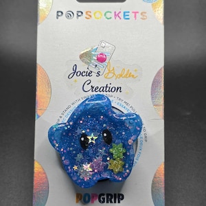 May include: A blue star-shaped PopSocket with glitter and star accents. The PopSocket has a cartoon face and is attached to a white and holographic backing with the text "POPSOCKETS" and "Jocie's Golden Creation".