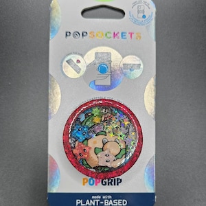 Puede incluir: Un PopSockets PopGrip en un paquete. El agarre redondo presenta un diseño colorido con un oso y otros personajes. El PopGrip es rojo con un efecto de brillo holográfico. El embalaje dice "POPSOCKETS" y "POPGRIP".