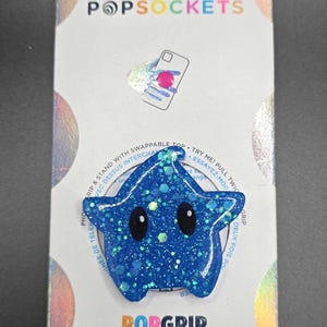 Op de afbeelding: Een blauwe, stervormige telefoonhouder met een glinsterende afwerking en twee zwarte ogen. De houder is verpakt op een witte kaart met de tekst "POPSOCKETS" en "POPGRIP" erop gedrukt.