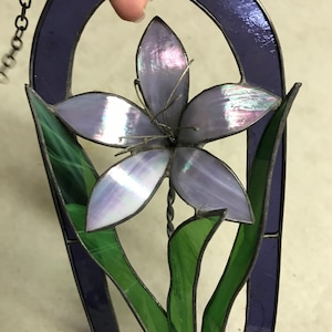 May include: A stained glass window hanging with a purple flower and green leaves in an oval frame.