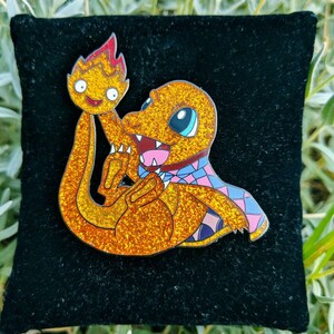 May include: A glittery orange enamel pin featuring a Charmander character holding a flame. The pin has a black background and is set against a backdrop of green foliage. The Charmander has blue eyes and a pink and blue patterned scarf.