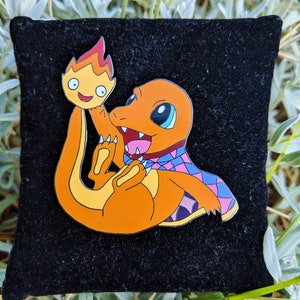May include: Orange enamel pin featuring a Charmander character with a flame. The pin has a black background and is set against a backdrop of green foliage. The Charmander is holding a flame.