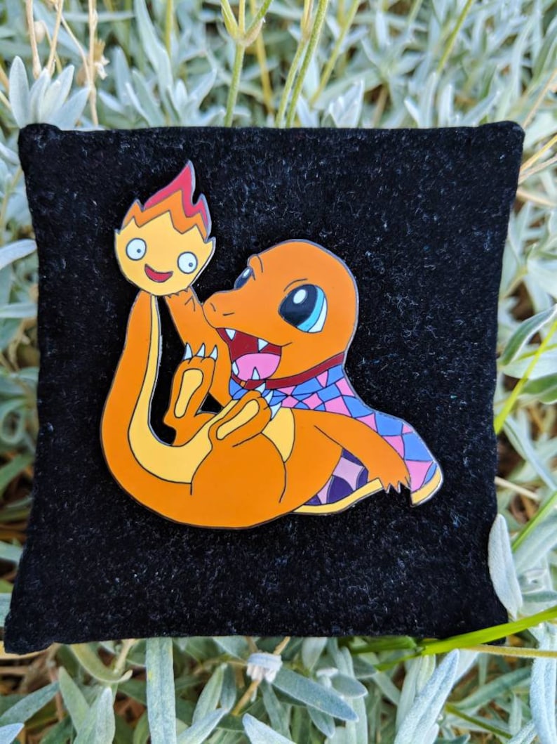 May include: Orange enamel pin featuring a Charmander character with a flame. The pin has a black background and is set against a backdrop of green foliage. The Charmander is holding a flame.