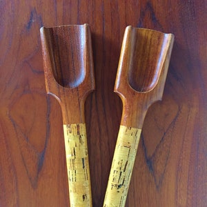 May include: Two wooden salad servers with cork handles. The servers are brown and have a scoop shape. The handles are a light brown color with a cork texture.