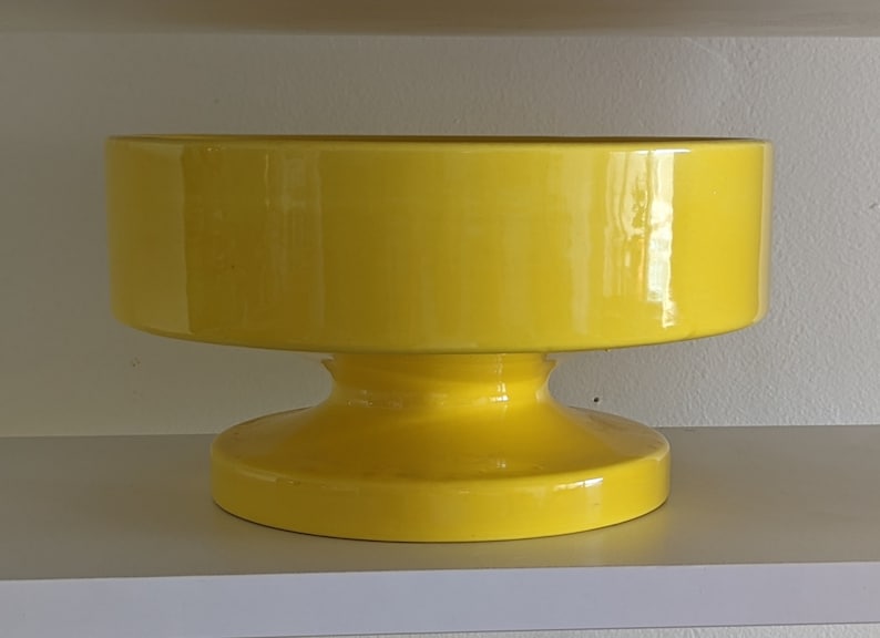 May include: A bright yellow ceramic bowl with a wide, round base. The bowl is on a white shelf.