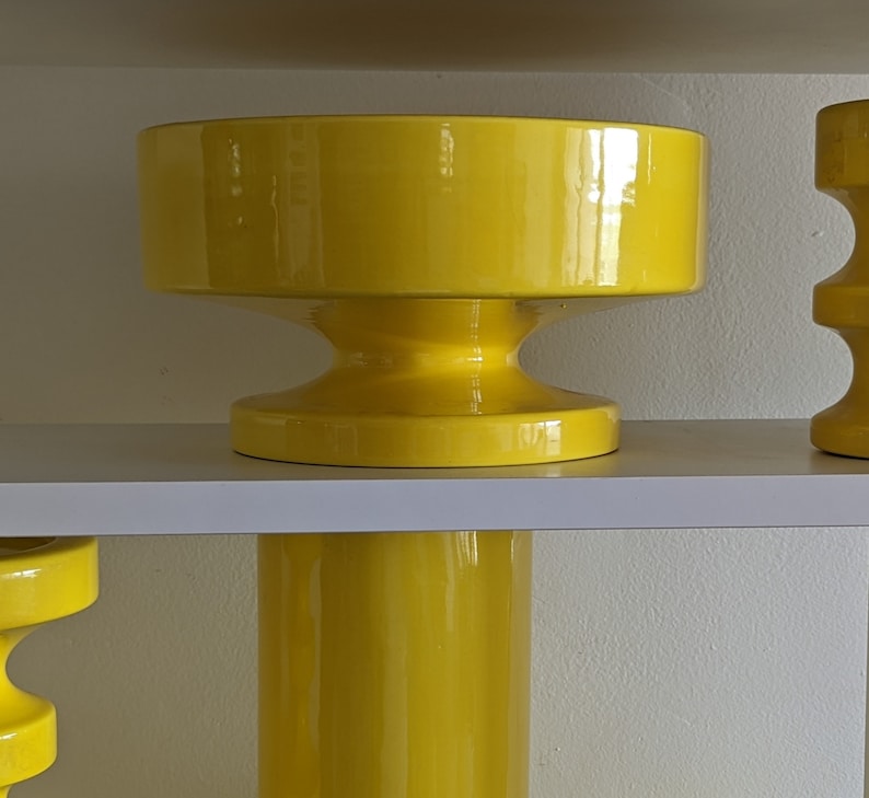 May include: A set of three yellow ceramic planters with a modern design. The planters are stacked on a white shelf.