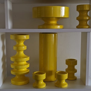 May include: A set of six yellow ceramic vases of varying sizes and shapes, arranged on a white shelf. The vases are all in a simple, modern style.