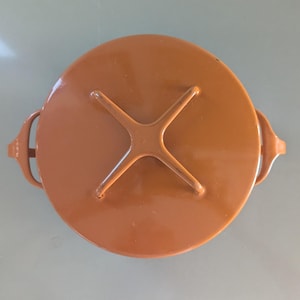 May include: A round, brown lid with a cross-shaped handle in the center and two side handles. The lid is made of a solid, opaque material and has a smooth surface. The lid is likely for a pot or container.