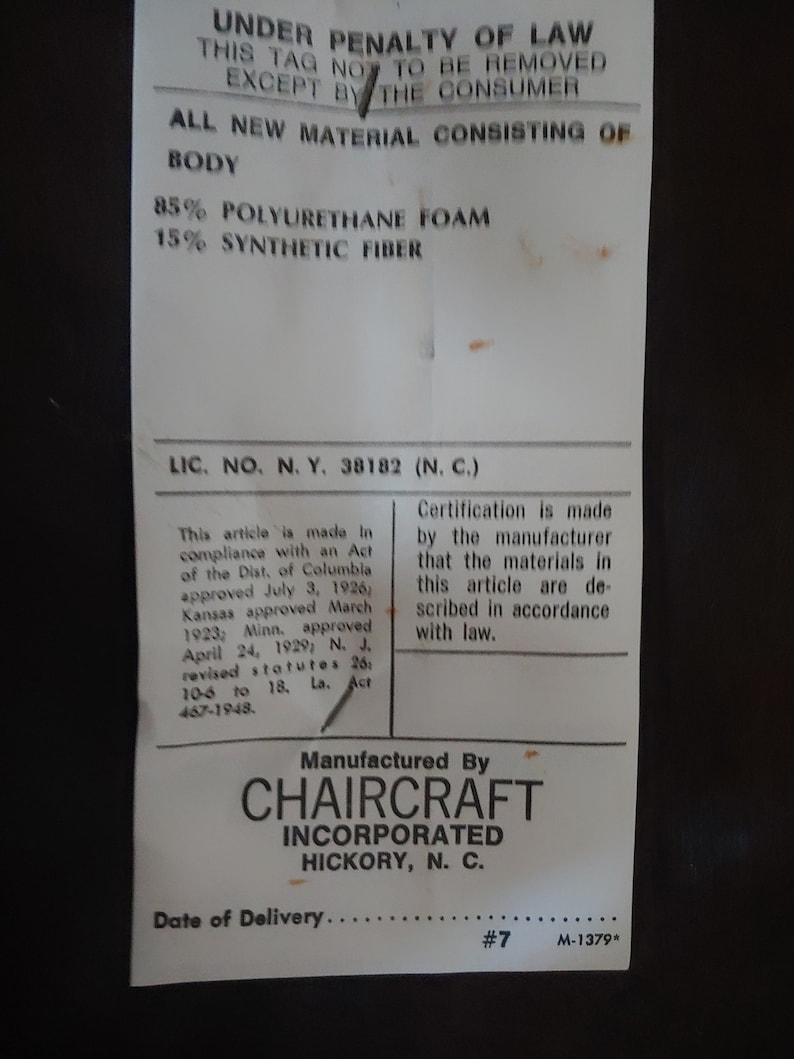 May include: A white tag with black text stating "Under Penalty of Law This Tag Not To Be Removed Except By The Consumer" and "All New Material Consisting of Body 85% Polyurethane Foam 15% Synthetic Fiber". The tag also includes the text "Lic. No. N.Y. 38182 (N.C.)" and "Manufactured By CHAIRCRAFT INCORPORATED HICKORY, N.C.". The tag has a handwritten number "7" and the text "M-1379*" at the bottom.