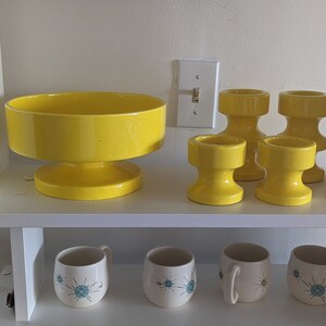 May include: A set of four yellow ceramic candle holders and a large yellow ceramic bowl with a pedestal base. The candle holders are all the same size and shape, with a cylindrical base and a flared top. The bowl is larger and has a wider base. All of the pieces are in a mid-century modern style.