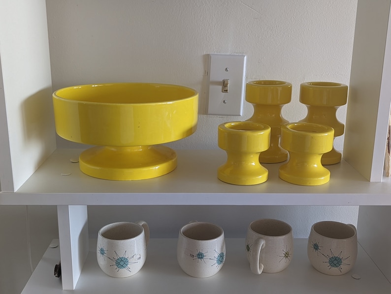 May include: A set of four yellow ceramic candle holders and a large yellow ceramic bowl with a pedestal base. The candle holders are all the same size and shape, with a cylindrical base and a flared top. The bowl is larger and has a wider base. All of the pieces are in a mid-century modern style.