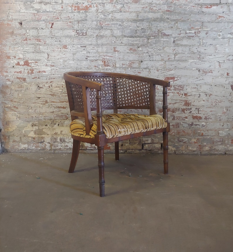 May include: A brown wooden chair with a woven cane back and seat. The chair has a yellow and brown striped fabric cushion. The chair is sitting on a concrete floor in front of a brick wall.
