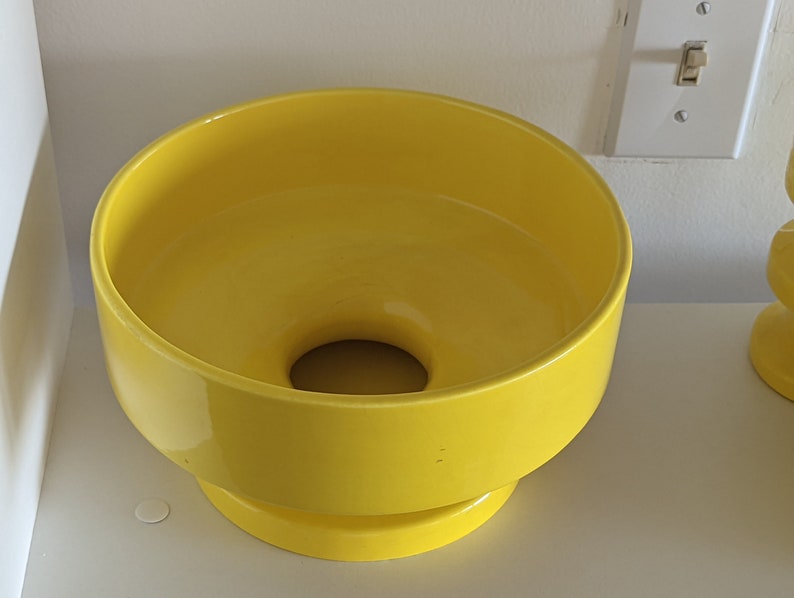 May include: A bright yellow ceramic planter with a wide, round opening and a smaller, round base. The planter is made of a smooth, glossy ceramic.