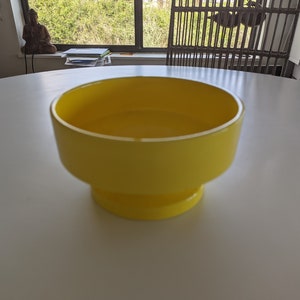 May include: A bright yellow ceramic bowl with a wide, round opening and a flared base. The bowl is sitting on a white surface.