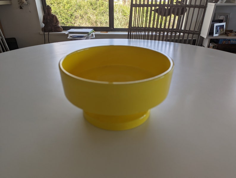 May include: A bright yellow ceramic bowl with a wide, round opening and a flared base. The bowl is sitting on a white surface.