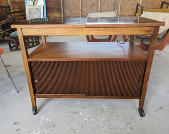 May include: A mid-century modern wooden serving cart with a black top, two shelves, and sliding doors. The cart has a dark wood finish and is on wheels.