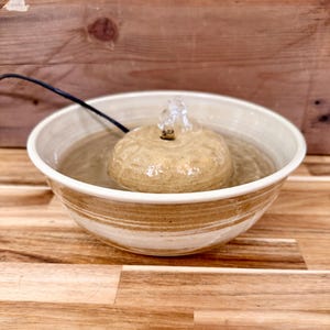 以下が含まれることがあります： クリーム色とタン色のストライプが入った広いボウルの中に、ベージュの質感のある丸いドーム型のセンターピースを備えたセラミック製のペット用給水器。ドームの頂上から小さな水流が湧き出ており、黒い電源コードが中央から伸びています。
