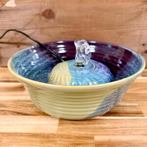 Könnte beinhalten: Ein Keramik-Trinkbrunnen für Haustiere mit einer breiten, gerippten Schale, die eine mehrfarbige Glasur in Blau-, Lila- und Gelbtönen aufweist. Ein kuppelförmiges Mittelstück sitzt in der Schale und stößt über ein schwarzes Kabel Wasser aus.