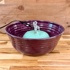 May include: A deep purple ceramic fountain bowl with a light teal fountain base. A clear, decorative water feature is in the center. The bowl has a ridged texture. A black power cord is visible.