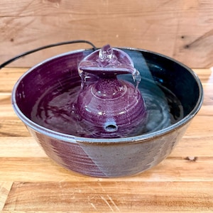 May include: A ceramic pet water fountain featuring a deep purple and dark blue glazed finish. The fountain has a central tiered structure with a small spout at the top where water flows, and a circular basin filled with water and a black power cord.