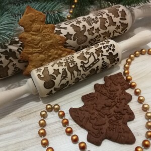 May include: Two wooden rolling pins with Christmas-themed designs, one with a snowman and the other with a reindeer, are shown with two gingerbread cookies shaped like Christmas trees. The cookies are decorated with the same designs as the rolling pins.
