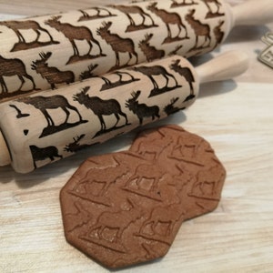 Puede incluir: Dos rodillos de amasar de madera con un diseño de alce grabado en ellos. Los rodillos de amasar están sobre una superficie de madera con una galleta marrón estampada con el diseño del alce.