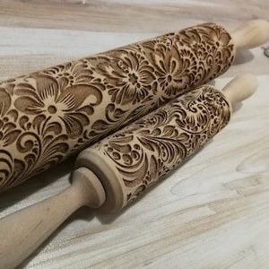 May include: Two wooden rolling pins with intricate floral designs carved into the surface. The rolling pins are made of natural wood and have a light brown color. The larger rolling pin has a handle on one end and the smaller rolling pin has a handle on both ends.