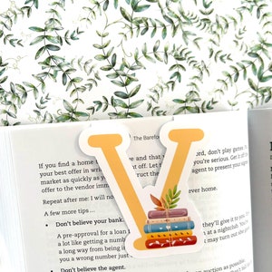 May include: A yellow letter V bookmark featuring a stack of colourful books and autumn leaves. The bookmark is placed on a page of a book with text that reads "Don't believe your bank." and "Don't believe the agent."