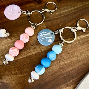 Peut inclure: Deux porte-clés avec des brins perlés ronds dans des tons roses et bleus. Chaque porte-clés comporte un charme circulaire avec une lettre et un motif de cœur. Les perles sont dans différentes nuances de rose, blanc et bleu, avec des accents argentés.