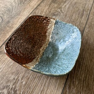 Może przedstawiać: Kwadratowy ceramiczny talerz o teksturowanej powierzchni. Talerz podzielony jest na dwie części: jedna jest brązowa, a druga niebieska. Obręcz ma ciemnobrązowy kolor. Talerz umieszczony jest na drewnianej powierzchni.