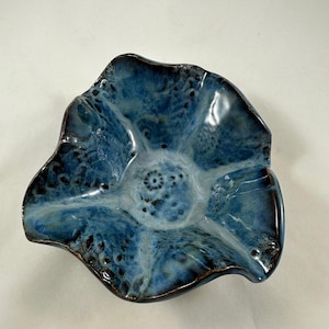 Puede incluir: Un cuenco de cerámica hecho a mano con una forma orgánica única. El cuenco presenta un esmalte azul moteado brillante con acentos más oscuros alrededor de los bordes y detalles interiores. El borde del cuenco tiene un ribete marrón oscuro.