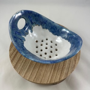 May include: A handmade ceramic colander with a white interior and a blue, textured rim. The colander has a series of small holes for draining and a single handle hole. It sits on a round, wooden base.