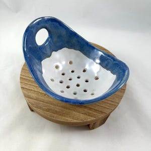 May include: A ceramic berry bowl featuring a glossy white interior with drainage holes and a vibrant blue glazed exterior rim. The bowl includes a single integrated handle with a circular cutout, providing a functional design for rinsing fruit.