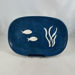 May include: A blue oval ceramic platter with a marine theme. Two white fish and white seaweed-like shapes are on the surface. The platter is displayed on a black metal stand.