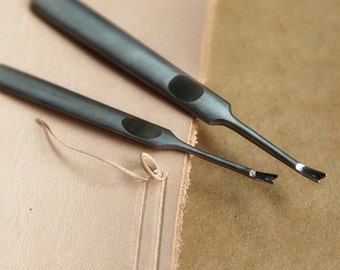 May include: Two silver leather crafting tools with black handles and pointed tips. The tools are lying on a light brown leather surface.