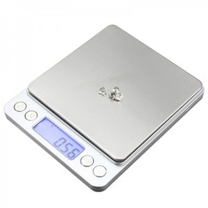 May include: A silver digital scale with a blue LCD screen displaying "0.56". The scale has four buttons on the front and a stainless steel weighing platform. Three small, clear gemstones are on the platform.