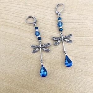 Op de afbeelding: Een paar oorhangers met een zilveren libel-bedel met heldere strass steentjes en blauwe kristallen kralen. De oorhangers hebben een traanvormige blauwe kristal aan de onderkant.